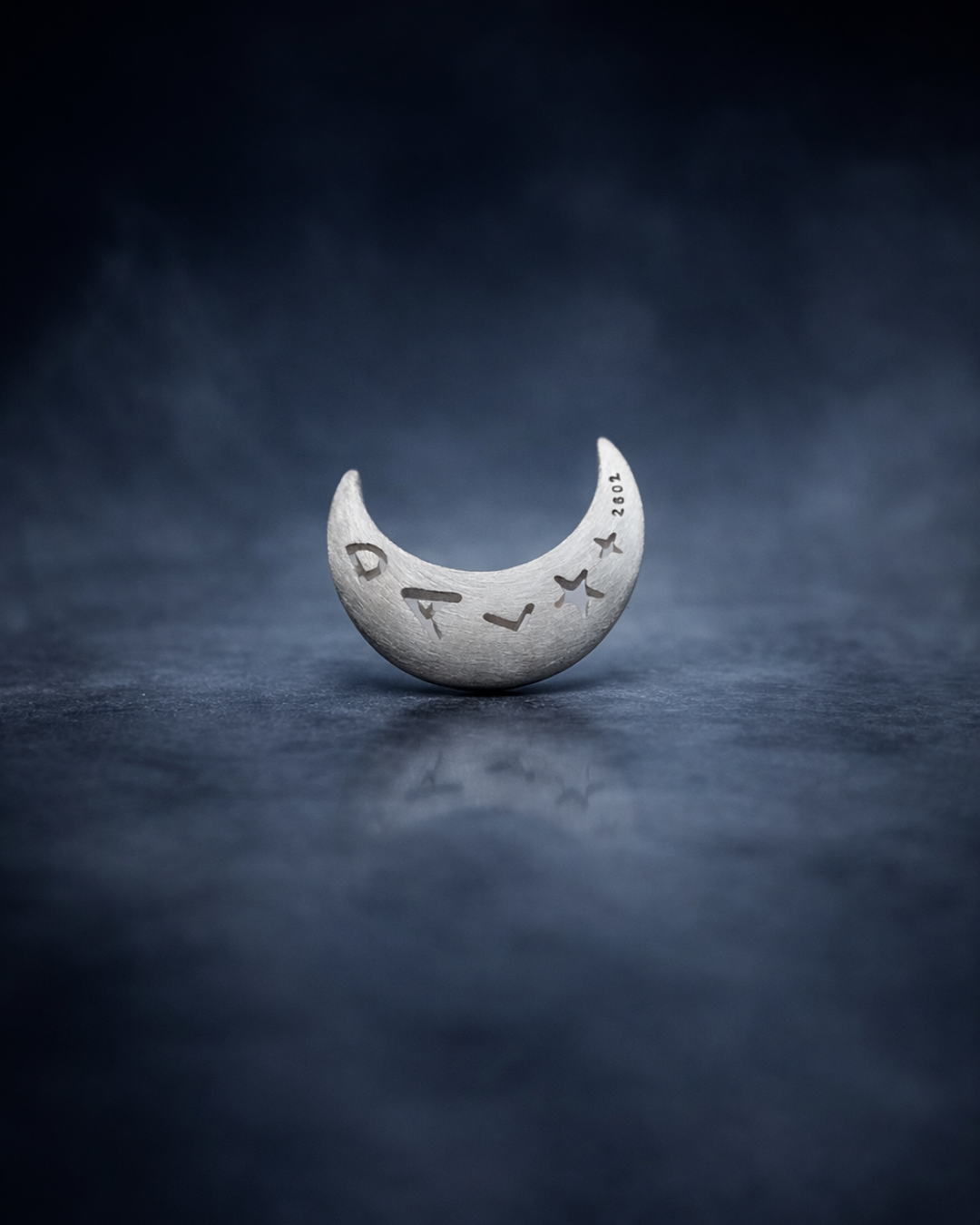 Silver crescent moon-shaped object with engraved text on a dark textured background